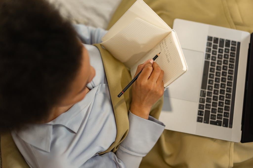 Overhead view of a woman taking notes in a notebook while using a laptop, embodying home learning.